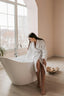 Woman in a white robe standing next to a freestanding bathtub in a bright room with large windows.